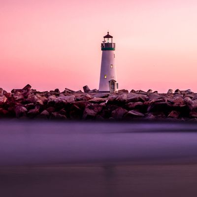 A lighthouse on a rocky shore, with purple water reflecting a rose sunset sky.
